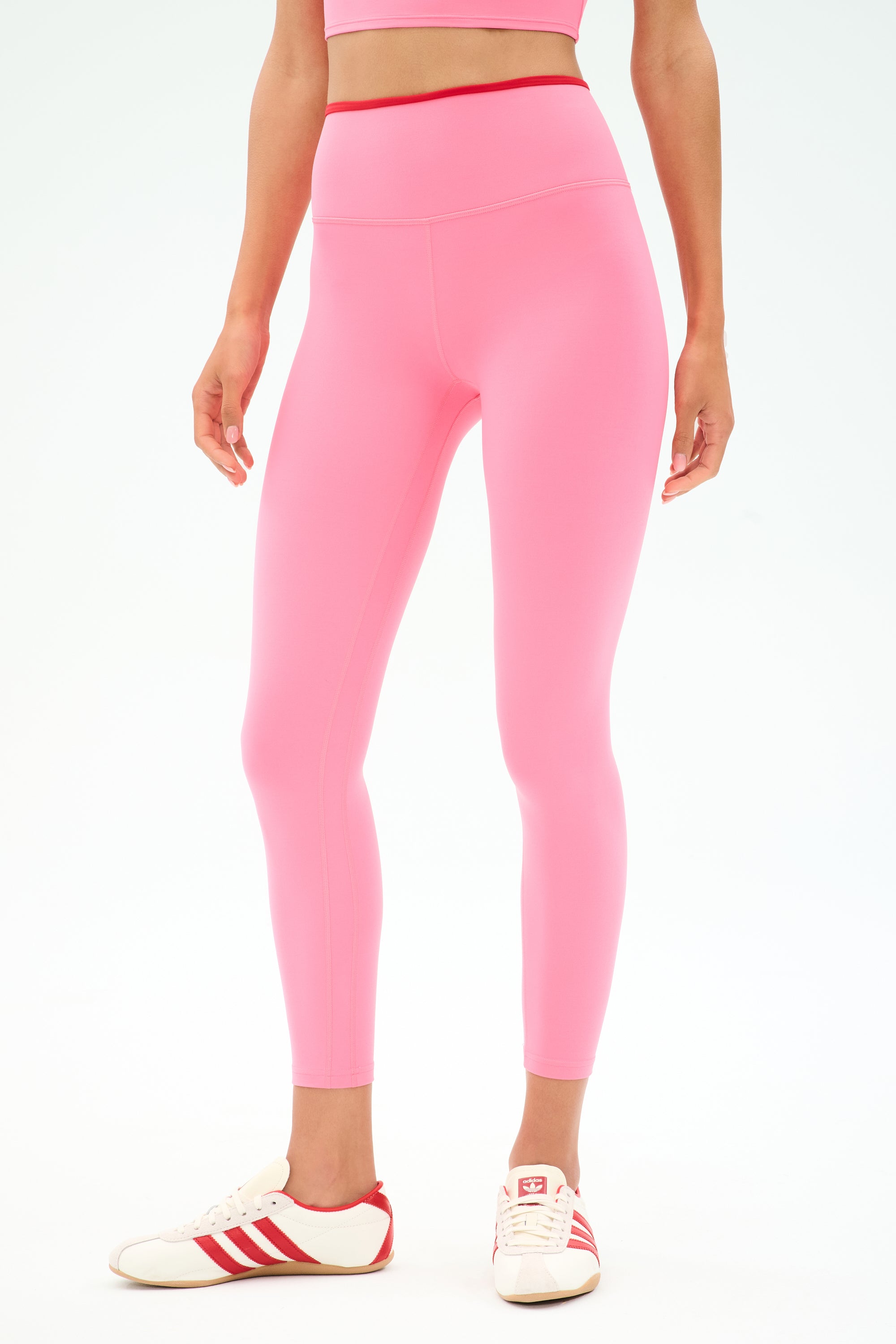 Wearing SPLITS59’s Easton Rigor High Waist 7/8 leggings in Strawberry/Pirate Red with a matching top and white sneakers with red stripes, standing against a plain light background—ideal for gym sessions or relaxed comfort.