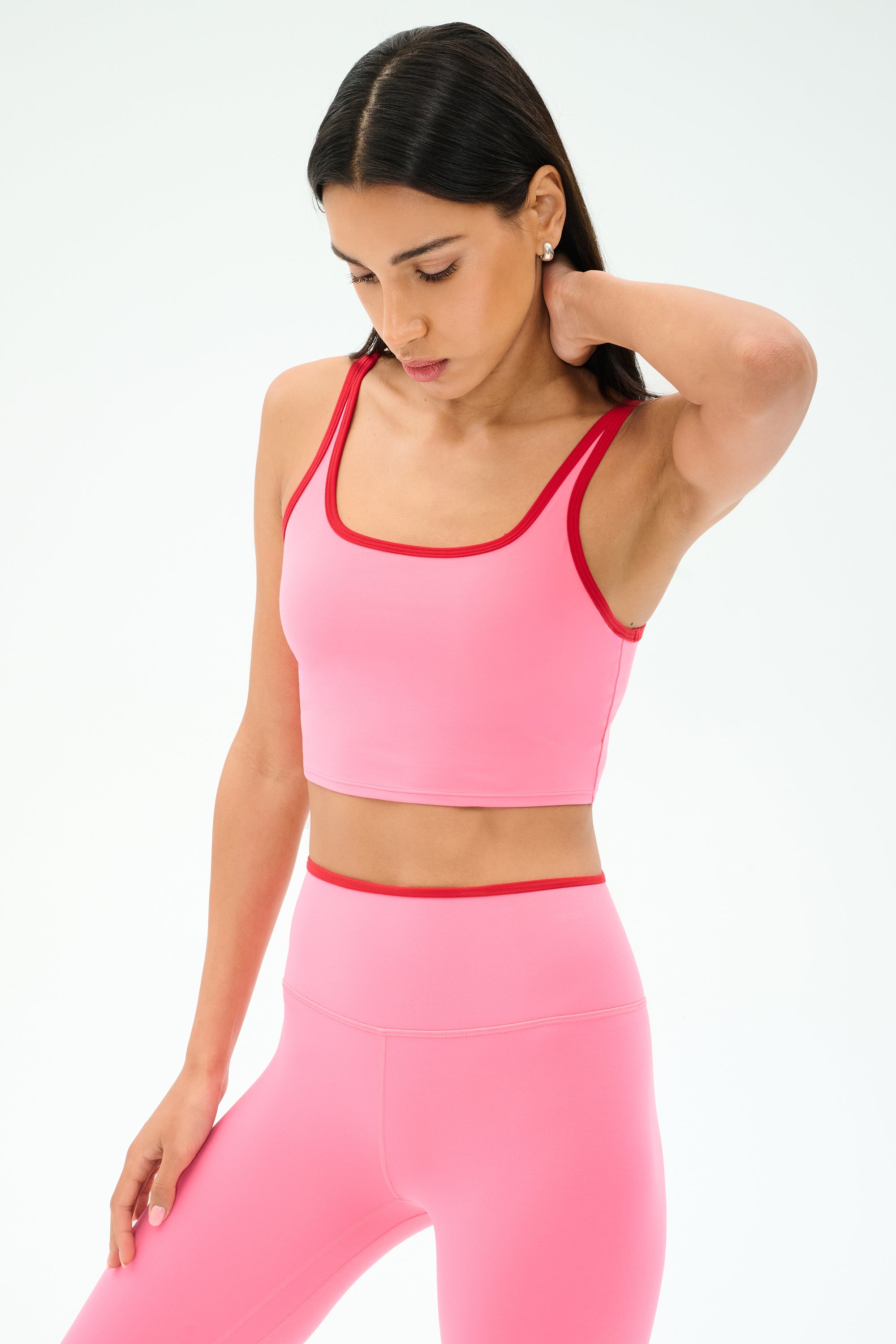 A woman wearing the SPLITS59 Cait Rigor Bralette in Strawberry/Pirate Red stands against a white background, resting one hand on her neck and looking down.