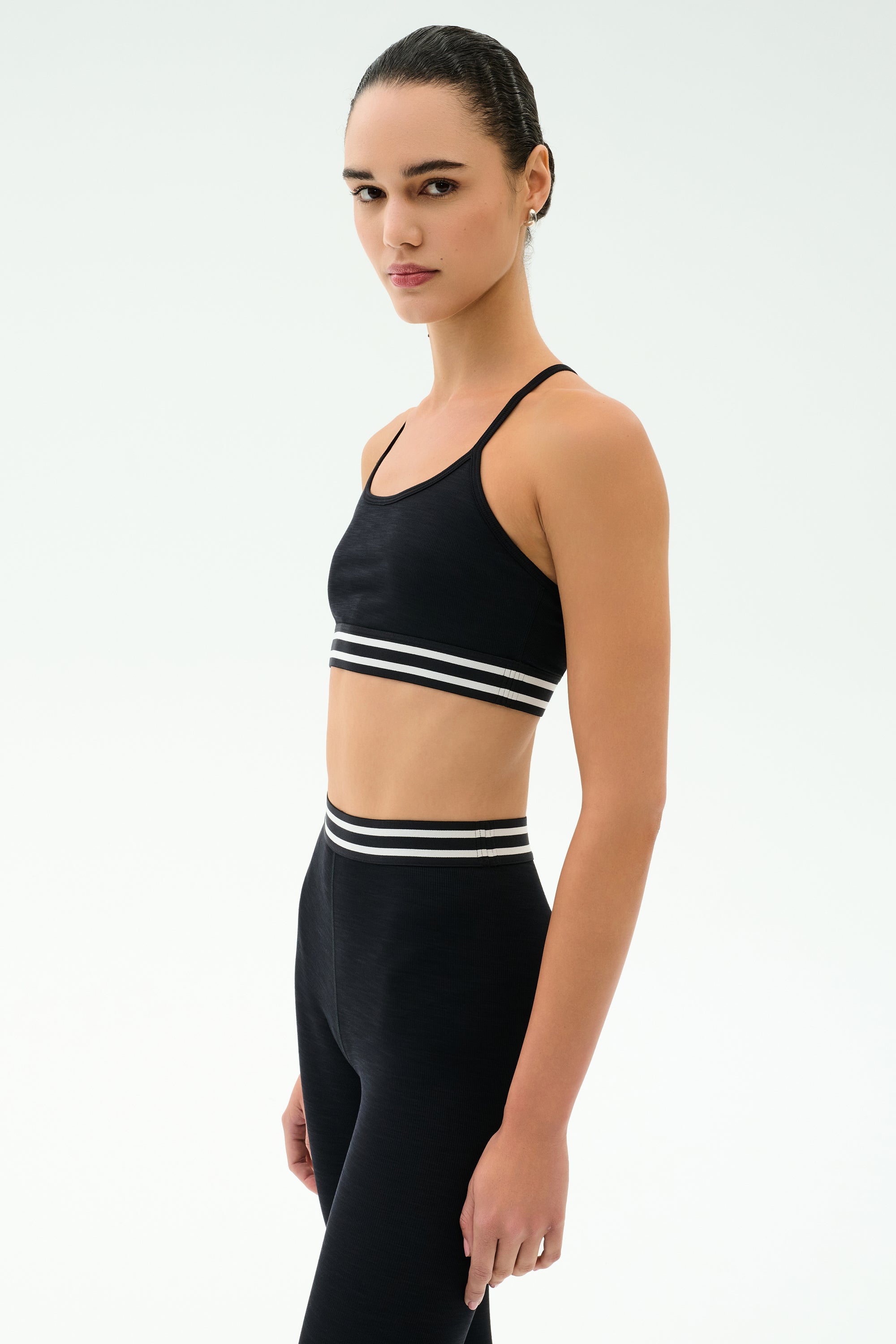 A woman stands sideways against a white background, wearing the SPLITS59 Cali Active Rib Bra in black and matching high-waisted leggings with white striped bands.