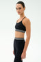 A woman stands sideways against a white background, wearing the SPLITS59 Cali Active Rib Bra in black and matching high-waisted leggings with white striped bands.