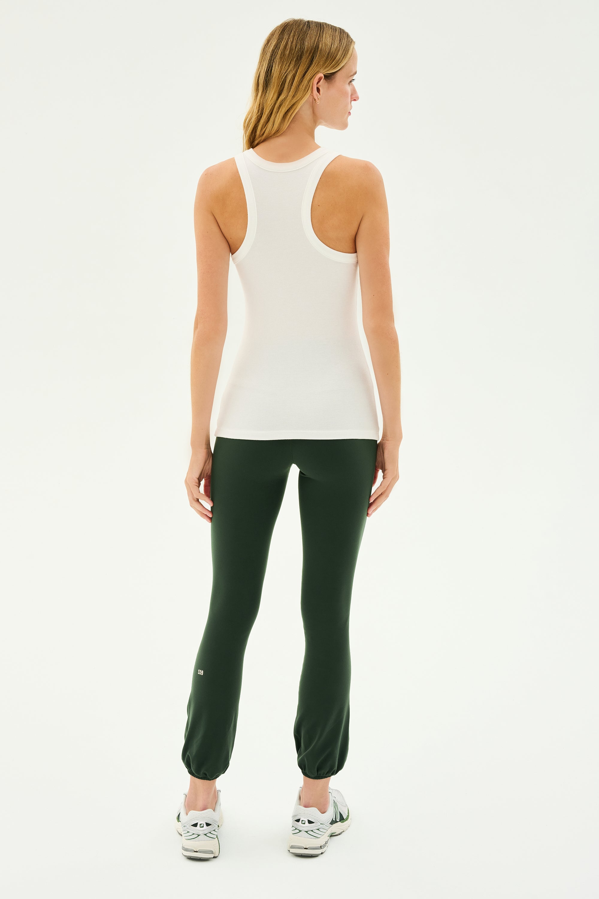 A woman stands facing away, dressed in a SPLITS59 Kiki Rib Tank Full Length in white, dark green leggings, and white sneakers, ready for some gym workouts.