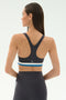 A woman faces away, wearing the SPLITS59 Maci Airweight Bra in Graphite/White, featuring a dark racerback design with blue, white, and gray bands, paired with matching dark leggings.