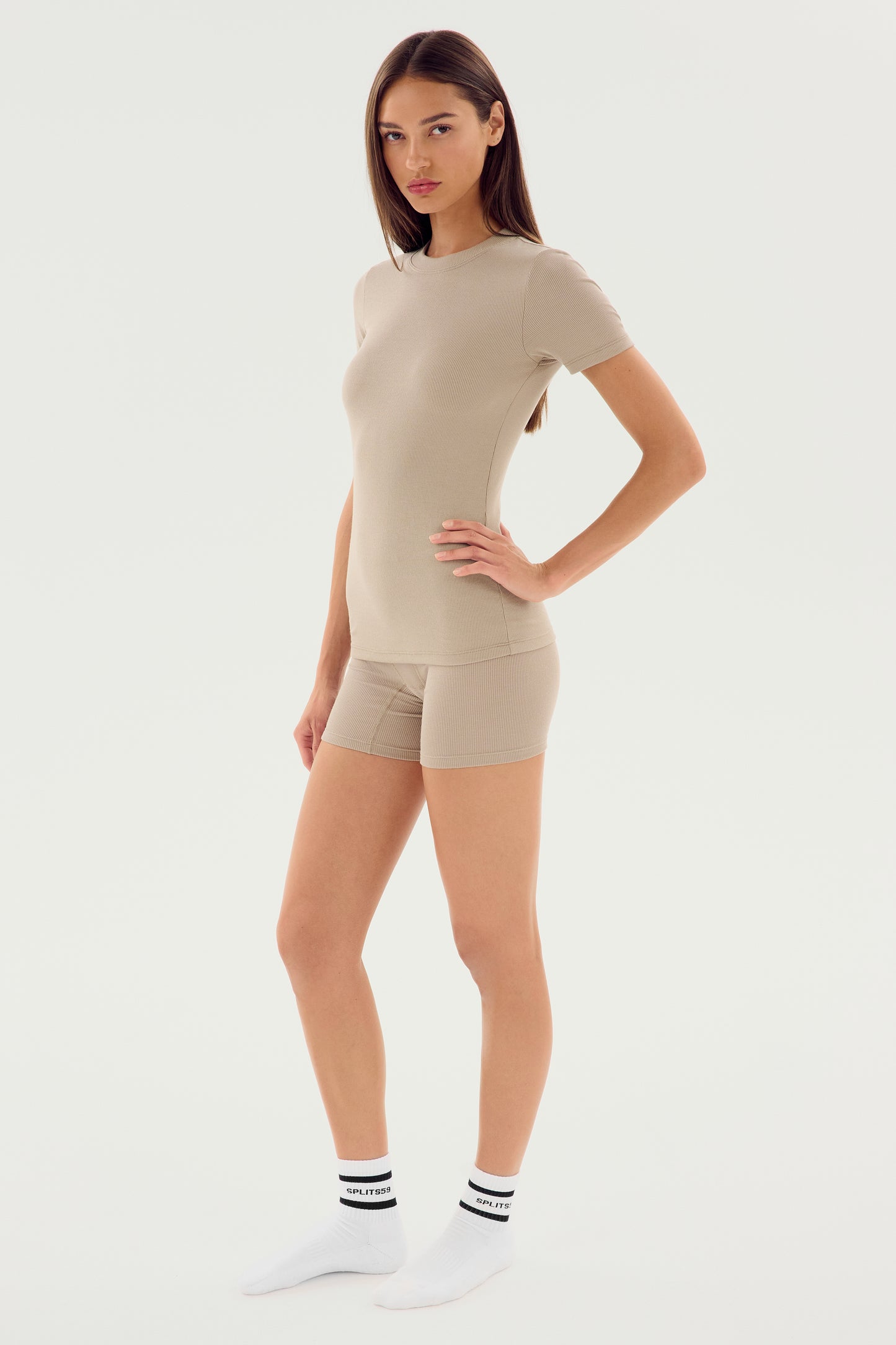 A woman stands against a plain background wearing the Louise Rib Short Sleeve in Latte by SPLITS59, paired with matching shorts and white socks.