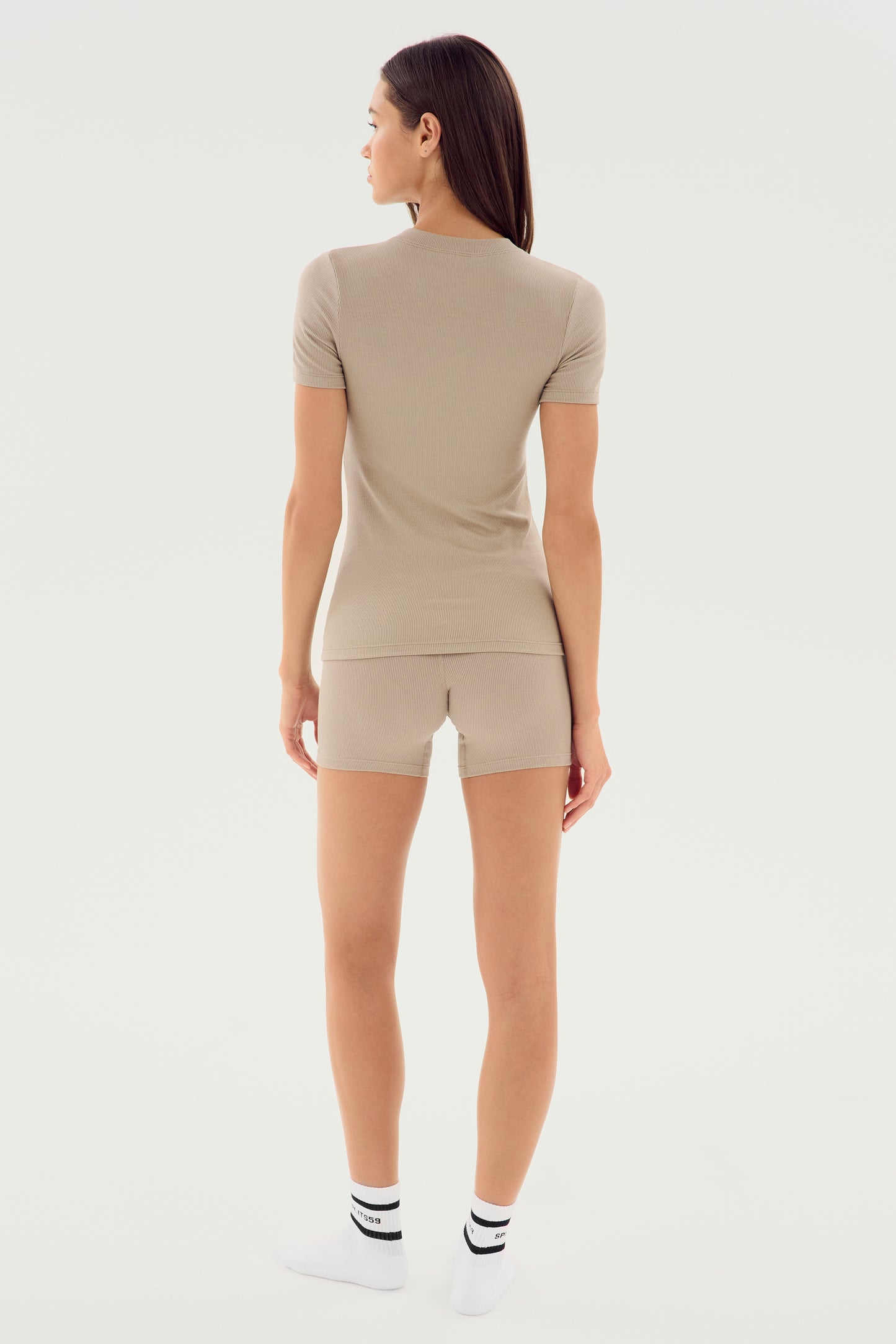 A person wearing the Louise Rib Short Sleeve - Latte from SPLITS59 and matching shorts, crafted from soft modal fabric, stands facing away, wearing white socks with black stripes against a plain white background.