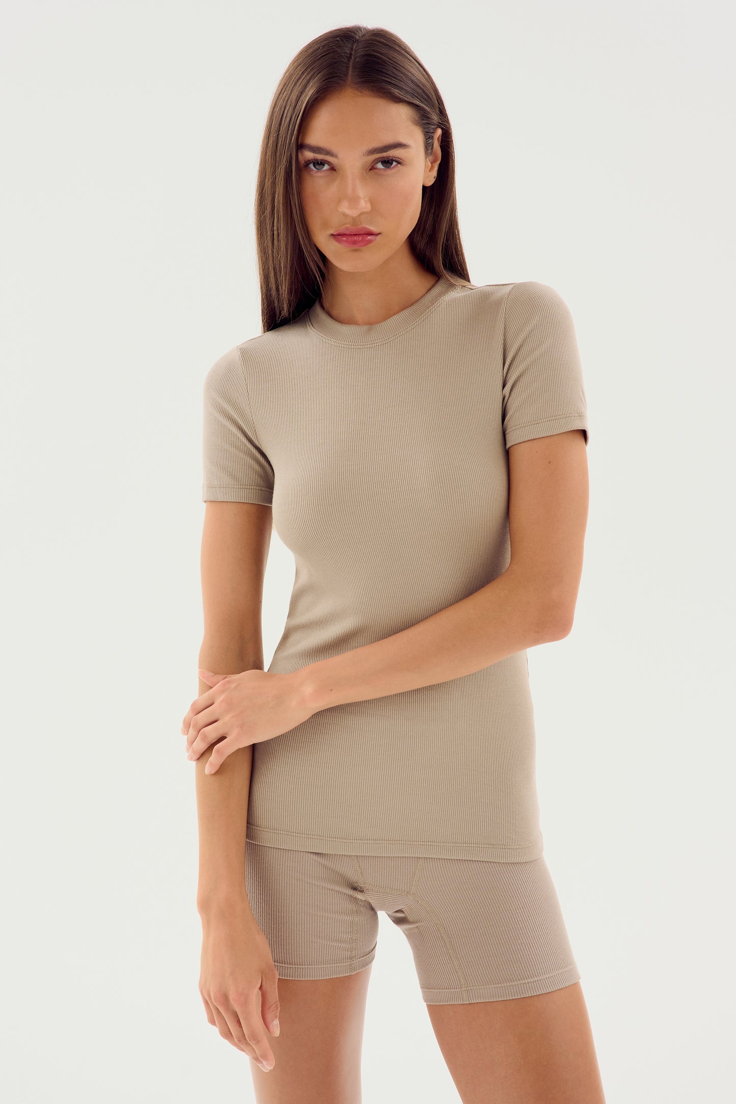 A woman with long brown hair, wearing the Louise Rib Short Sleeve in Latte by SPLITS59 along with matching shorts made from soft modal fabric, stands against a plain white background.