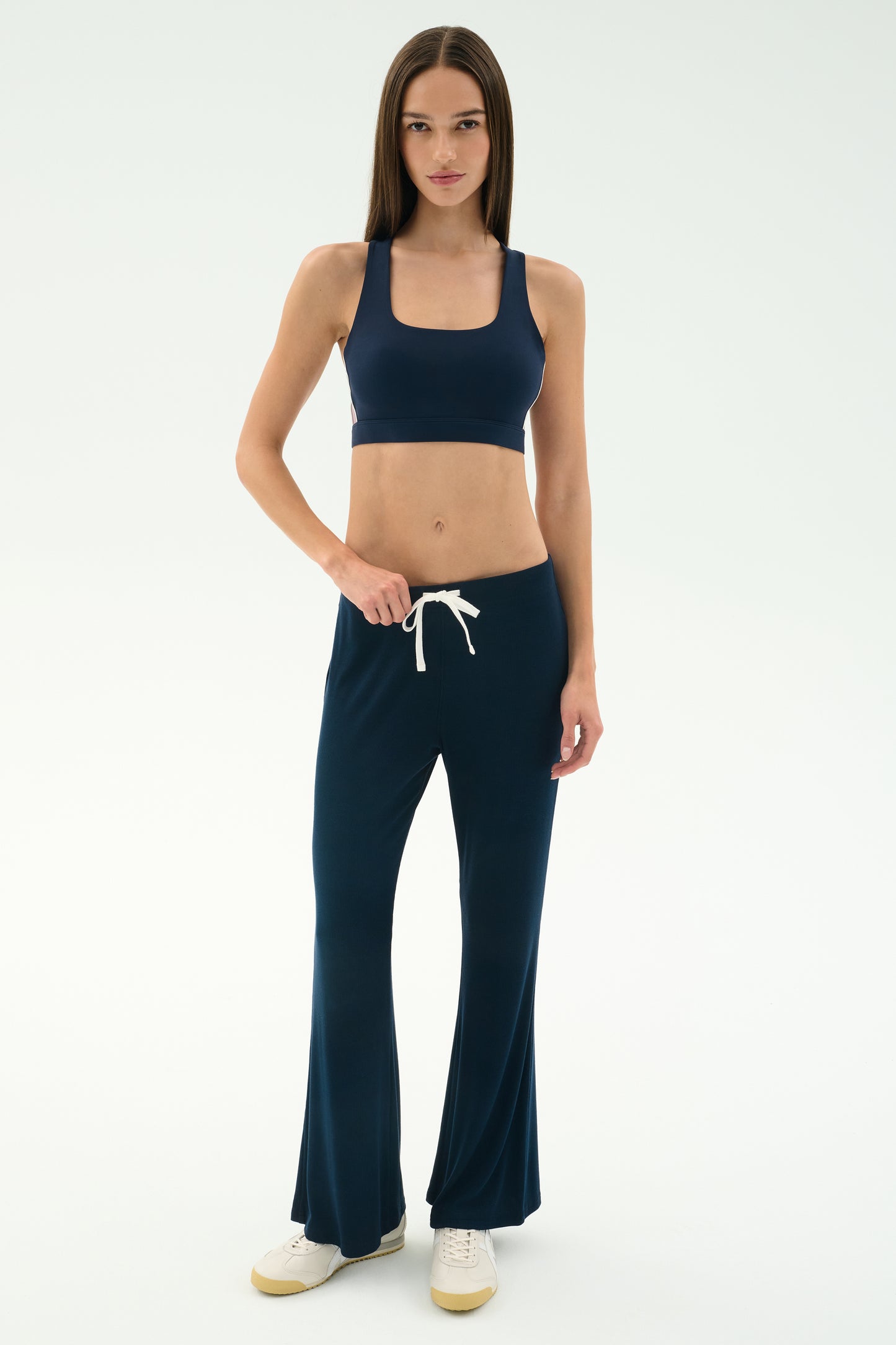 A woman stands against a plain background wearing the SPLITS59 Blaire Rib Pant in Indigo, a navy spandex sports bra, and white sneakers.