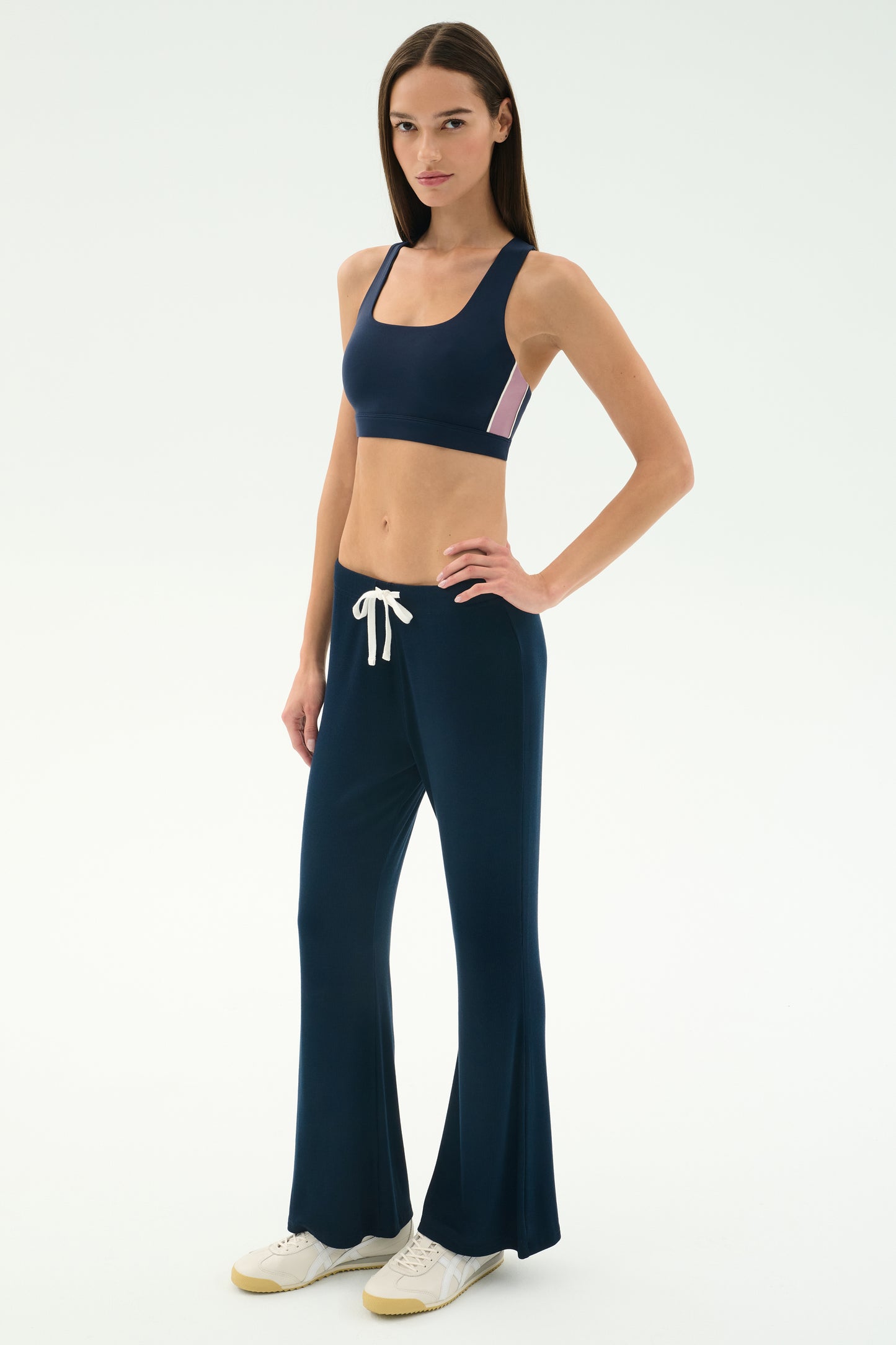 A woman wears the SPLITS59 Blaire Rib Pant in Indigo, made from soft modal and spandex, paired with a matching sports bra and white sneakers, posing against a plain white background.