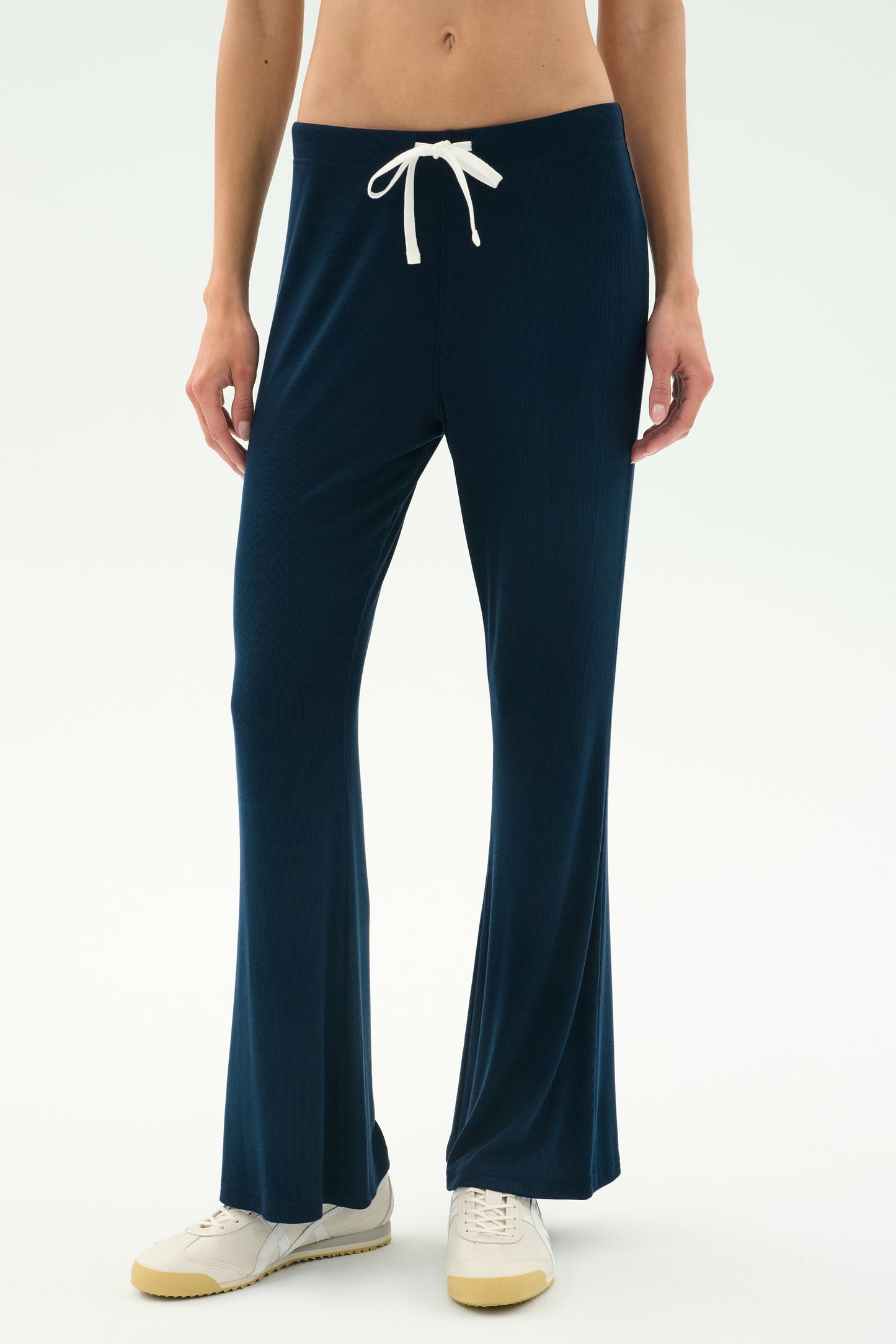 A person wears the SPLITS59 Blaire Rib Pant in Indigo—navy blue drawstring lounge pants crafted from soft modal with spandex—paired with white sneakers, standing against a plain light background.