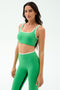 A woman wears the SPLITS59 Cait Rigor Bra in Grass/White, a green sports bra with white trim designed for high-impact workouts, paired with matching high-waisted leggings, standing against a plain white background.
