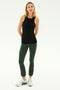 A woman stands against a plain background wearing the Kiki Rib Tank Full Length in black from SPLITS59, paired with dark green pants and white sneakers. She has her hands behind her back and looks straight ahead, embodying the calm focus of yoga.