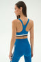 A woman stands facing away from the camera against a light background, wearing the SPLITS59 Maci Airweight Bra in Stone Blue/Graphite with racerback design, paired with matching blue leggings.
