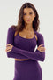 A woman with long brown hair is standing with her arms crossed, wearing a purple Shiri Airweight Shrug - Black Currant sports bra and matching high-waisted leggings made of sleek SPANDEX from SPLITS59.