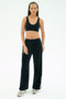 A woman stands against a white background wearing the SPLITS59 Airweight Scoop Back Bra in black, paired with black wide-leg pants and gray sneakers.