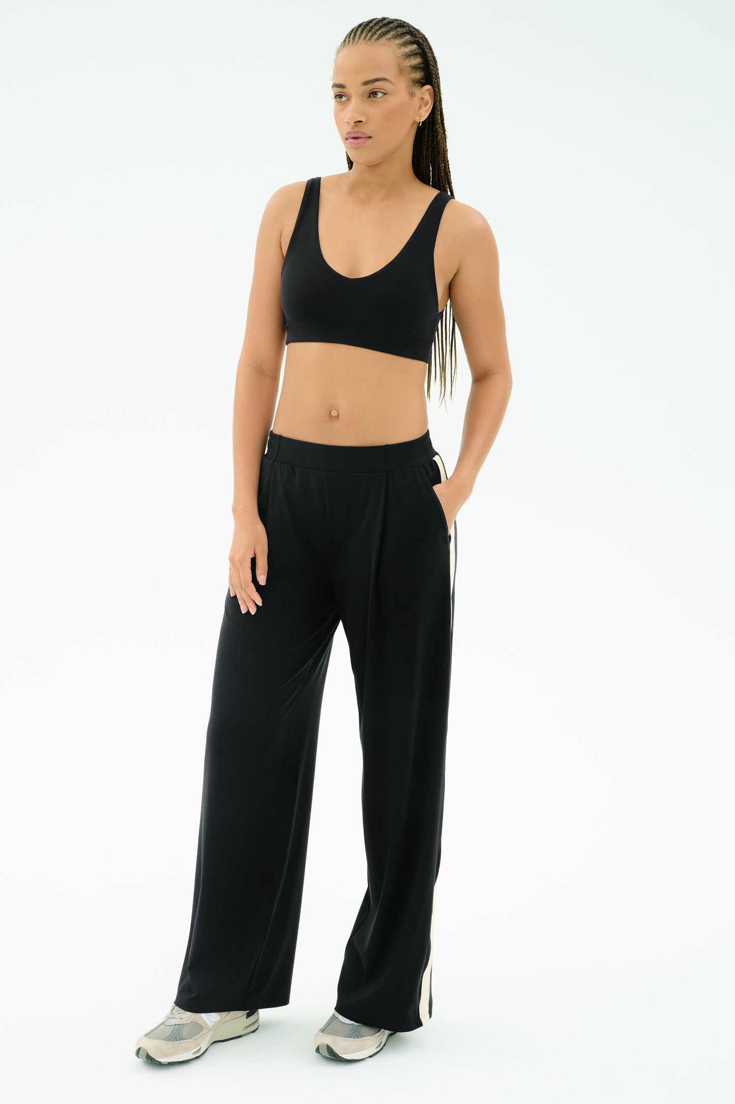 A woman poses against a plain background wearing the SPLITS59 Airweight Scoop Back Bra in black, paired with black wide-leg pants and sneakers, one hand in her pocket.