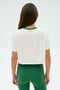 A person with long brown hair is seen from behind, wearing the SPLITS59 Djuna Crop Ringer in white/arugula, set against a simple, light backdrop.