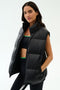 A woman stands against a plain light background wearing the SPLITS59 Summit Nylon Puffer Vest in black over a sports bra and leggings, ready for cool weather workouts with its lightweight insulation.