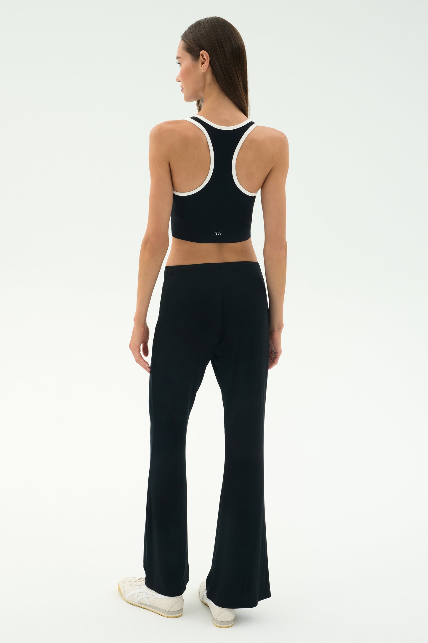 A woman faces away, wearing SPLITS59's Blaire Rib Pant in black with a black racerback crop top, styled with white sneakers against a plain light background.