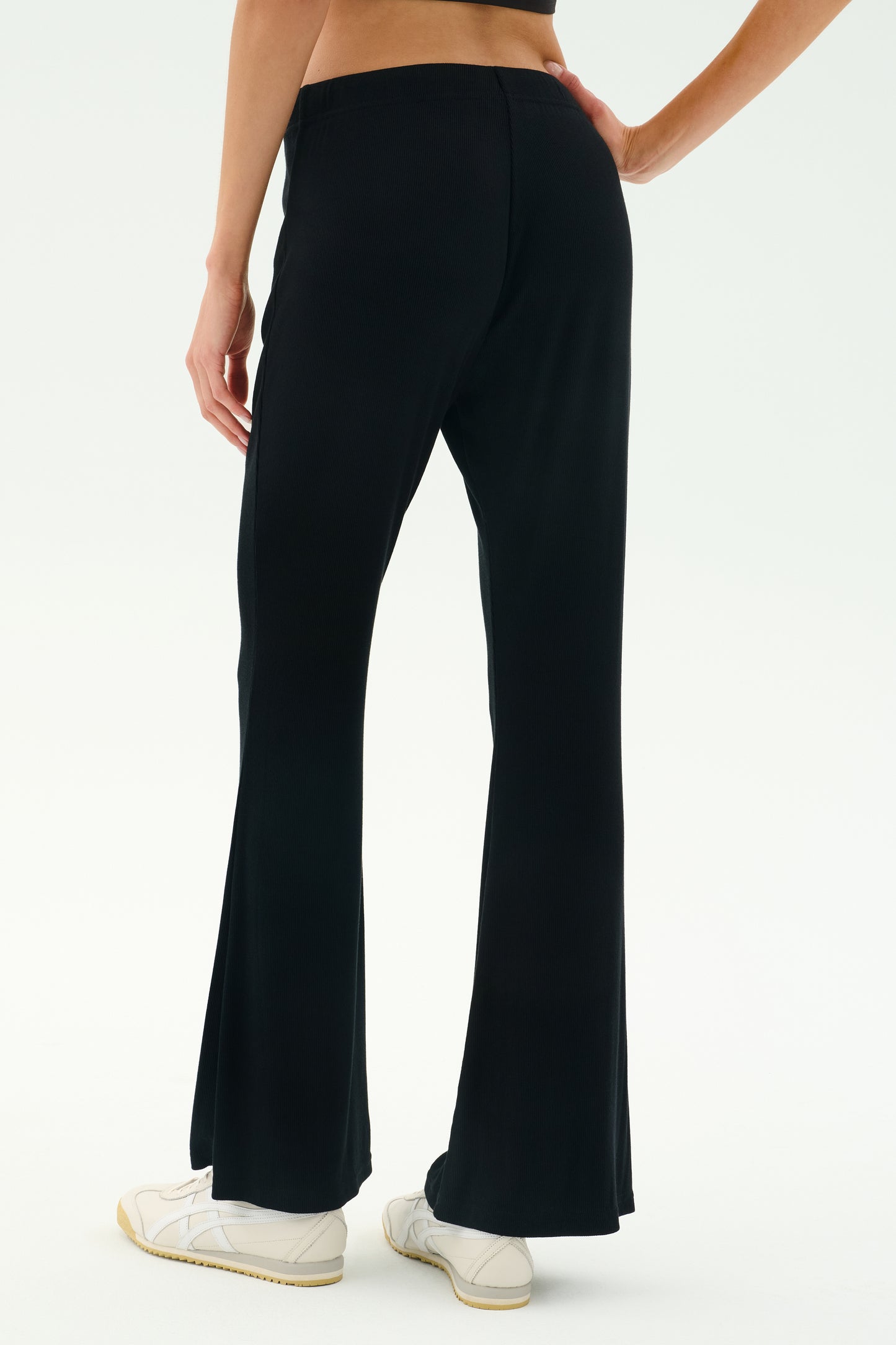 A person wearing SPLITS59 Blaire Rib Pant in black, made with soft modal and spandex, paired with white sneakers, stands on a white background, shown from the back waist down.