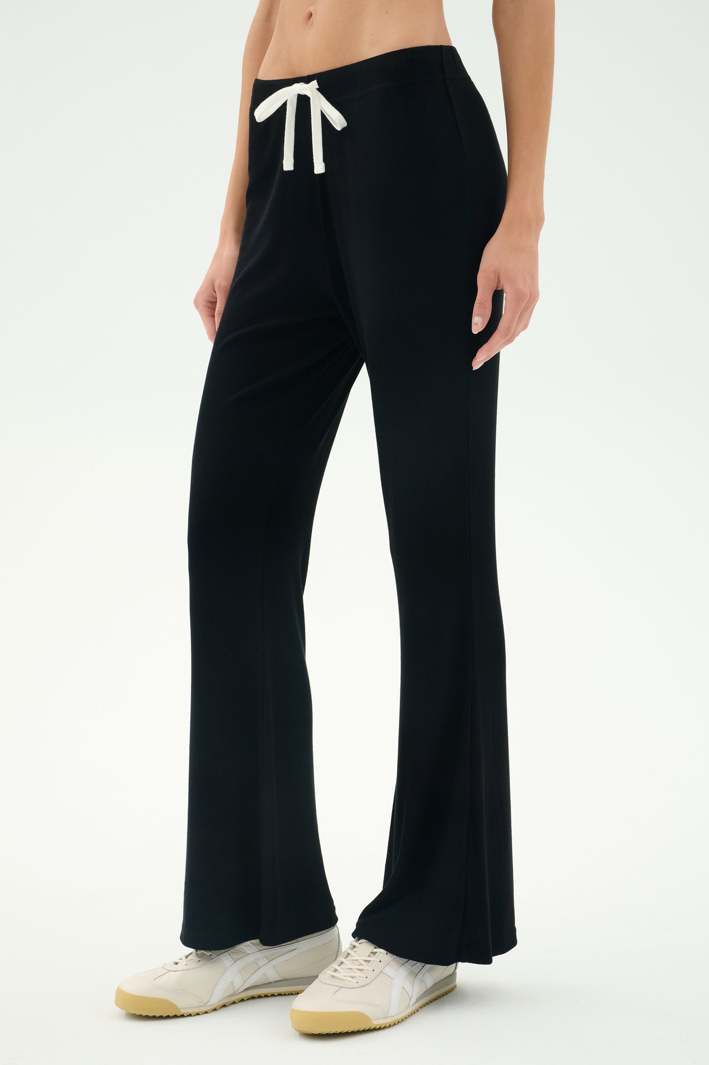 Someone wearing the SPLITS59 Blaire Rib Pant in black, which features a white drawstring, paired with white sneakers and standing against a plain light background.