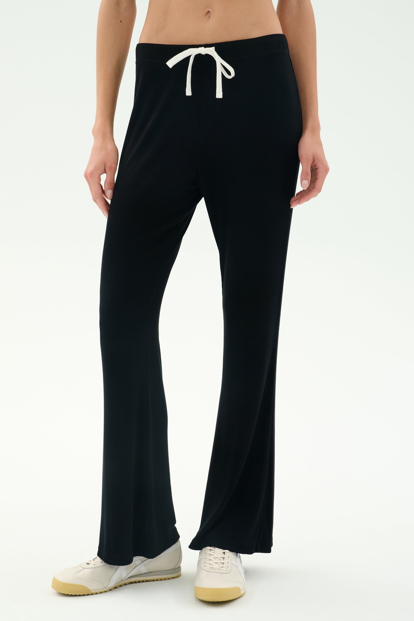 A person wears the SPLITS59 Blaire Rib Pant in black, featuring soft modal fabric, paired with white sneakers, standing against a plain white background.