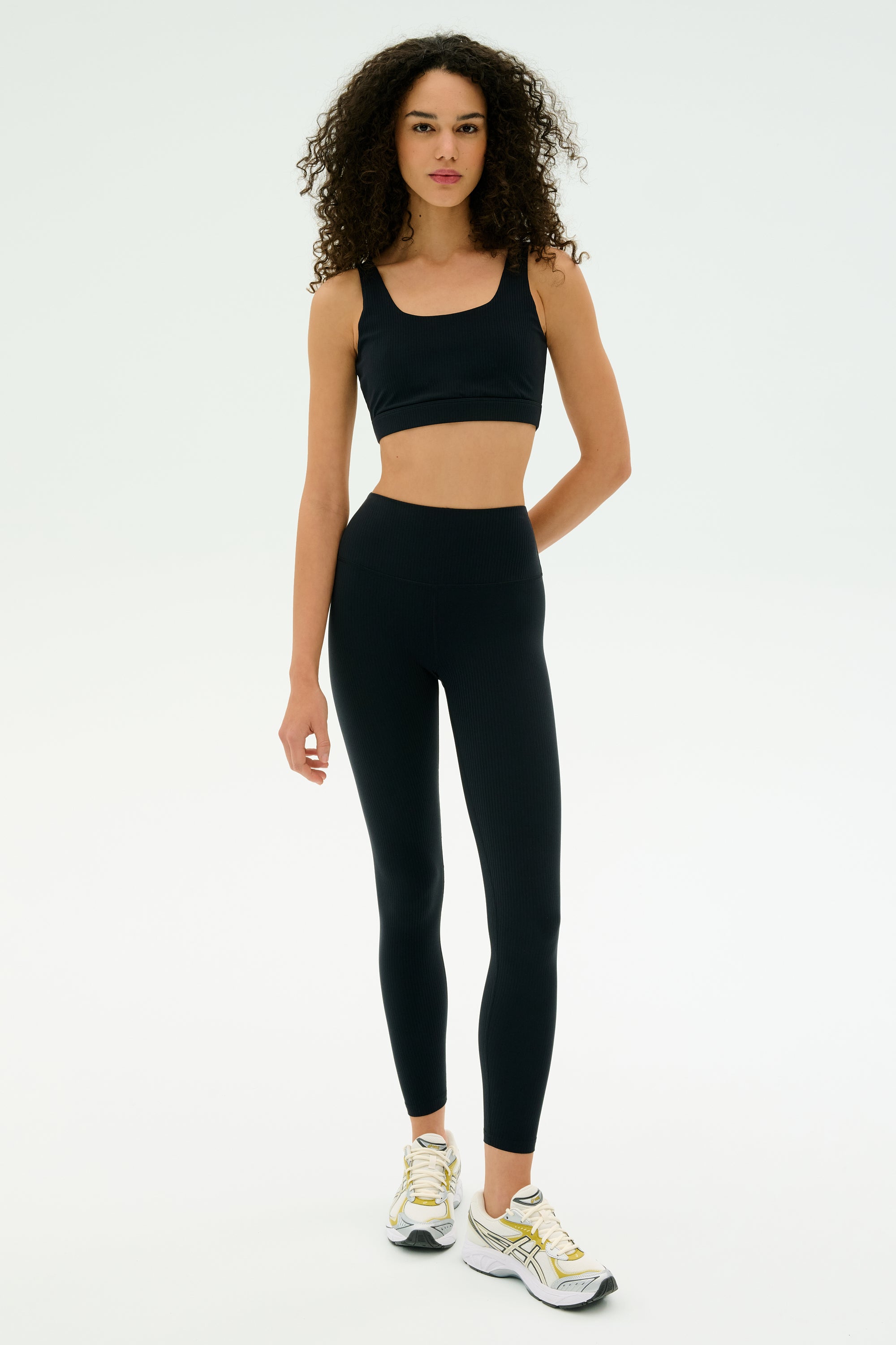 A woman stands against a plain background wearing the SPLITS59 Airweight Rib High Waist 7/8 leggings in black, paired with a matching black sports bra and white sneakers.