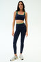 A woman faces forward in SPLITS59’s Easton Rigor High Waist 7/8 leggings and matching indigo/grass sports bra, made of compressive fabric, paired with white sneakers against a plain white background.