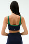 A woman with long dark hair wears the SPLITS59 Cait Rigor Bra in Indigo/Grass, featuring green trim and matching high-waisted leggings made from Rigor fabric, shown from the back against a plain light background.