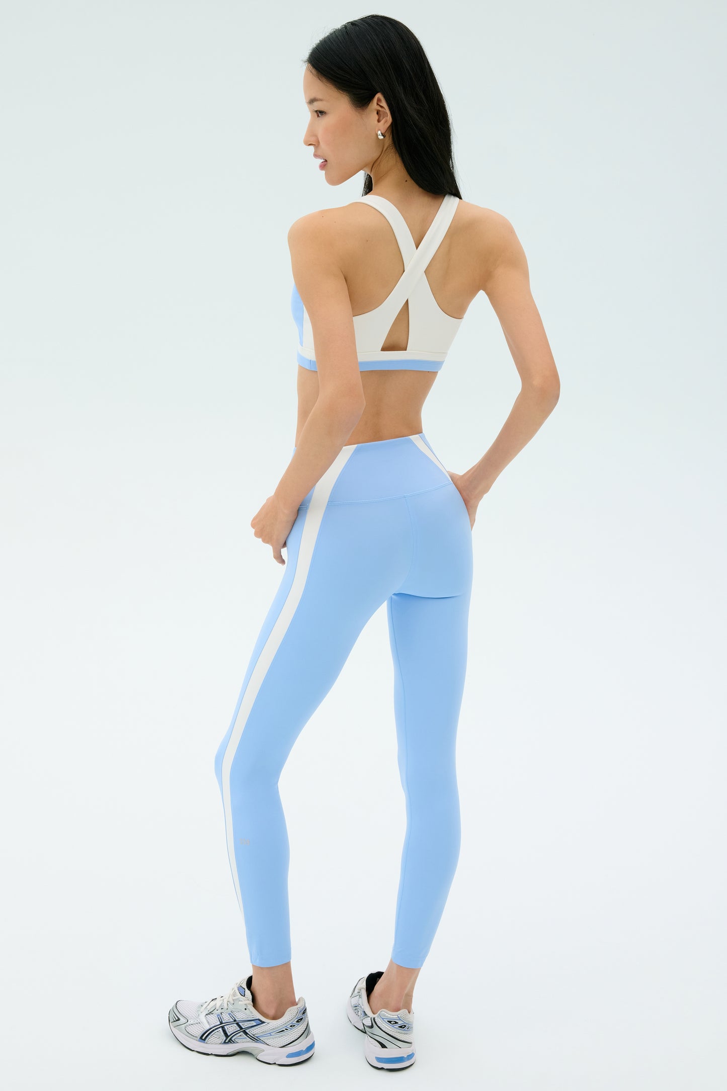 A woman wears the SPLITS59 Miles Rigor Bra in Skylight/White with matching leggings, hands on hips, standing slightly turned and wearing white running shoes.