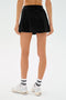 Model wears the SPLITS59 Venus High Waist Rigor Skort in black, paired with white socks featuring black stripes and white sneakers, shown from the back against a plain white background.