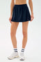 A person wears the SPLITS59 Venus High Waist Rigor Skort in black, paired with white ankle socks and sneakers, standing against a plain light background.