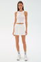 A woman stands against a white background wearing a sleeveless white knotted tank top, SPLITS59 Venus High Waist Rigor Skort in white, white sneakers, and white socks with black stripes.