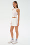 A woman stands against a plain light background wearing the SPLITS59 Venus High Waist Rigor Skort in white, a white sleeveless top with a front knot, white socks, and sneakers.