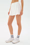 A person wears the SPLITS59 Venus High Waist Rigor Skort in white, paired with white sneakers and white socks with black lettering, standing against a plain white background.