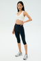 A woman stands against a plain background wearing the SPLITS59 Airweight Bra with Ruching in white, paired with black cropped leggings and silver running shoes.