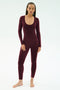 A person stands barefoot against a white background, wearing the SPLITS59 Chloe Airweight Jumpsuit in Dark Cherry—a fitted, long-sleeve piece with a scoop neckline made from smooth nylon spandex.
