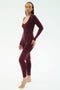 A person stands barefoot against a plain background, wearing the SPLITS59 Chloe Airweight Jumpsuit in Dark Cherry, a fitted long-sleeve piece made from a smooth nylon-spandex blend.