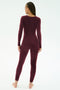 A person with braided hair stands barefoot in a well-lit studio, wearing the SPLITS59 Chloe Airweight Jumpsuit in Dark Cherry, featuring long sleeves and soft nylon spandex fabric.