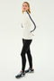 A person wearing a SPLITS59 Jackson Rib Full Length Turtleneck in White/Indigo with black stripes on the sleeves, paired with black leggings and black athletic shoes, stands against a plain white background—ready for a yoga or Pilates session.