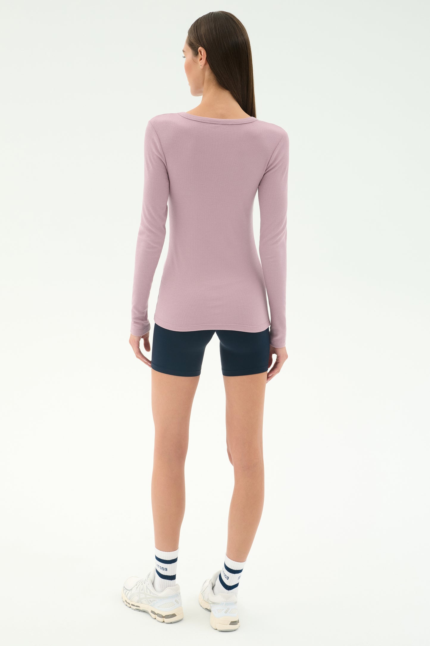 A woman stands facing away, wearing the SPLITS59 Louise Rib Henley in blush, navy blue shorts, white striped socks, and white sneakers against a plain white background.