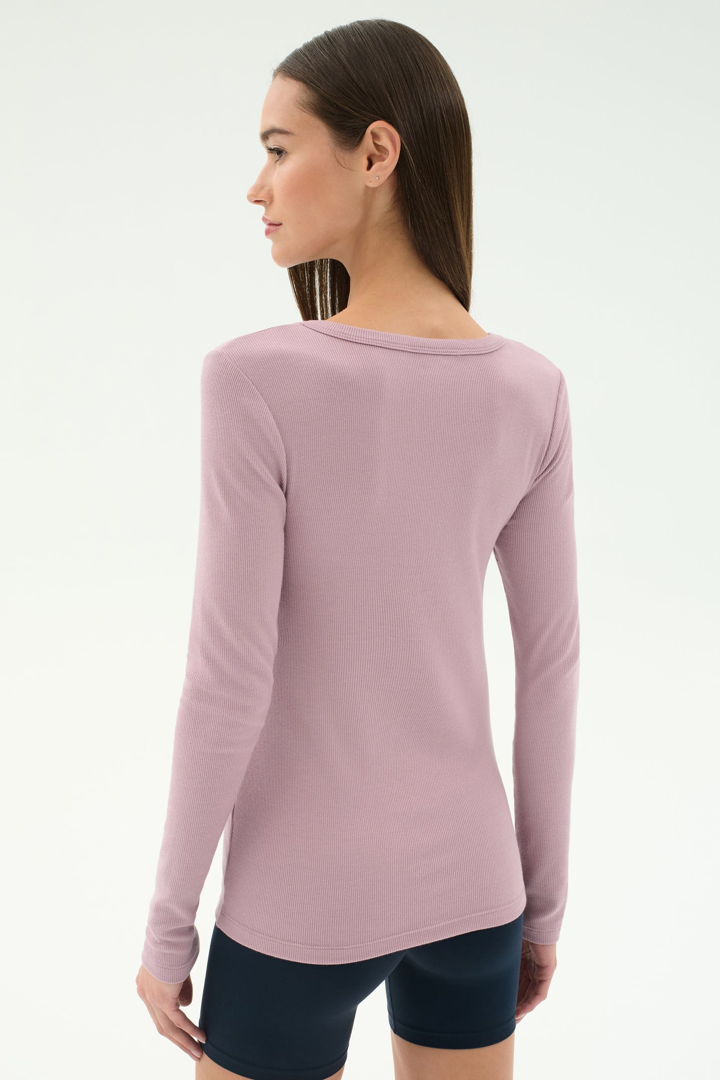 A woman with straight brown hair, facing away from the camera, wears the SPLITS59 Louise Rib Henley in blush and black shorts against a plain light background.