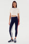 A woman wearing a white Djuna Crop Ringer - White/Indigo tee from SPLITS59, navy blue leggings with red and white side stripes, and black athletic shoes stands against a plain white background.