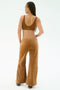A person with long braids stands facing away, wearing the SPLITS59 Joy Velour Pant in Honey—a pair of tan wide-leg pants made from soft polyester spandex—paired with a brown sports bra against a plain light background.