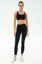A woman faces forward in a SPLITS59 Lexi Rigor Bra - Black/Dark Chocolate, styled with black high-waisted leggings and white sneakers, standing against a plain white background.