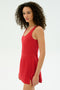 A woman with curly hair models the SPLITS59 Martina Rigor Dress in Pirate Red, sleeveless, standing against a plain white background and facing slightly to the side.