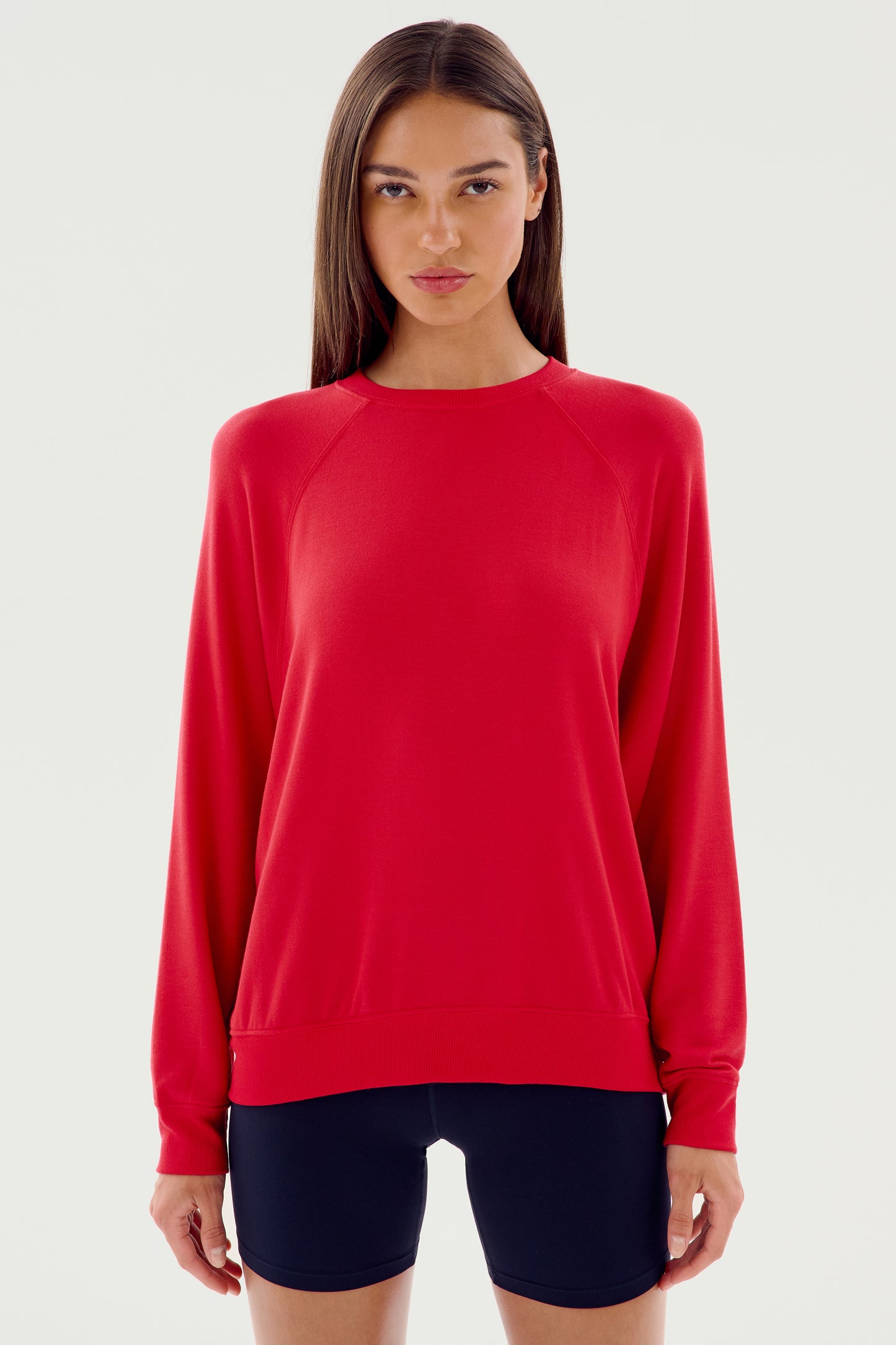 A person stands facing the camera wearing an Andie Fleece Sweatshirt - Pirate Red by SPLITS59 and black shorts against a plain white background.