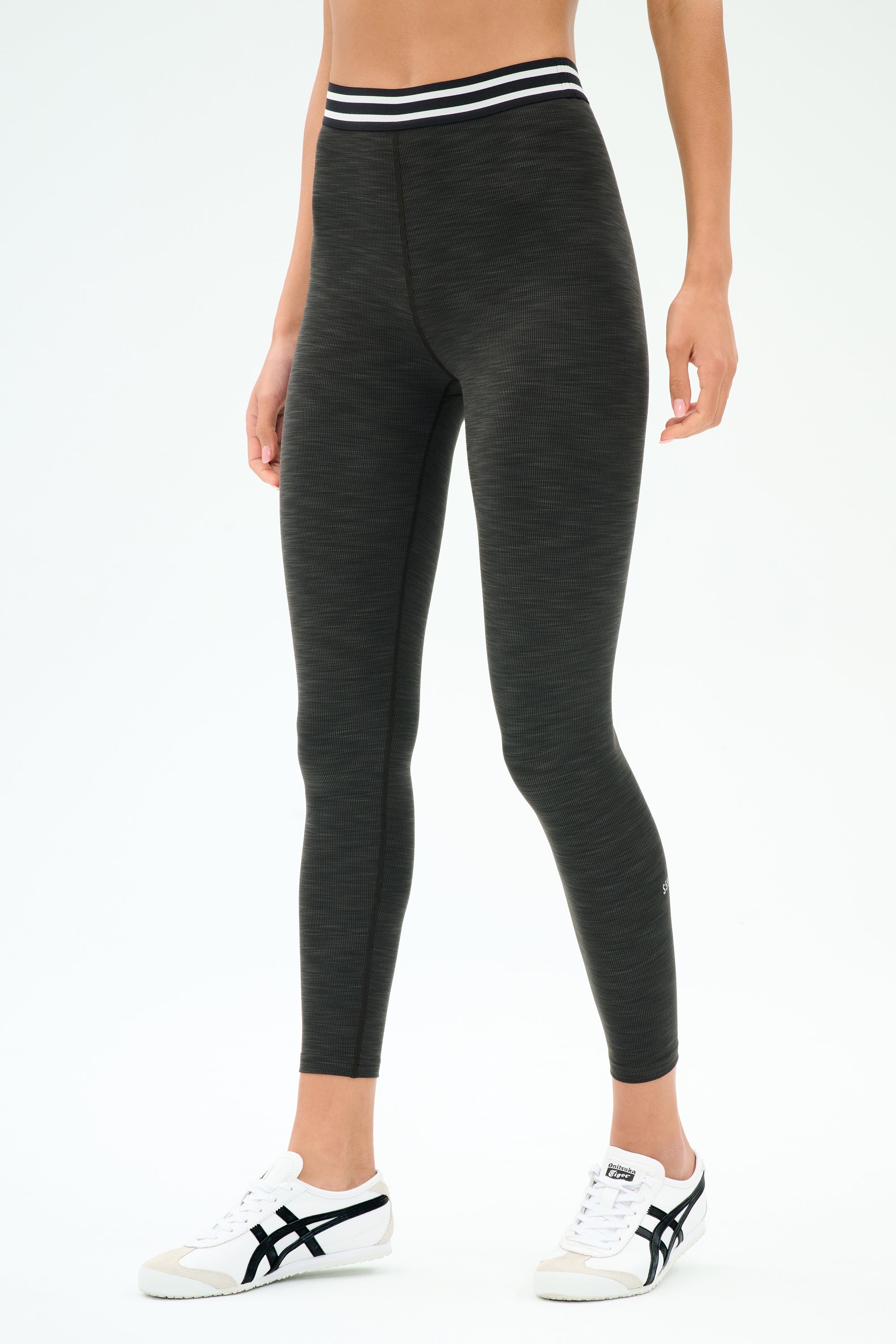 A person wears the SPLITS59 Bailey Active Rib High Waist 7/8 leggings in Heather Graphite, featuring a white striped waistband and white sneakers, standing against a plain background—ideal for gym workouts or casual wear.