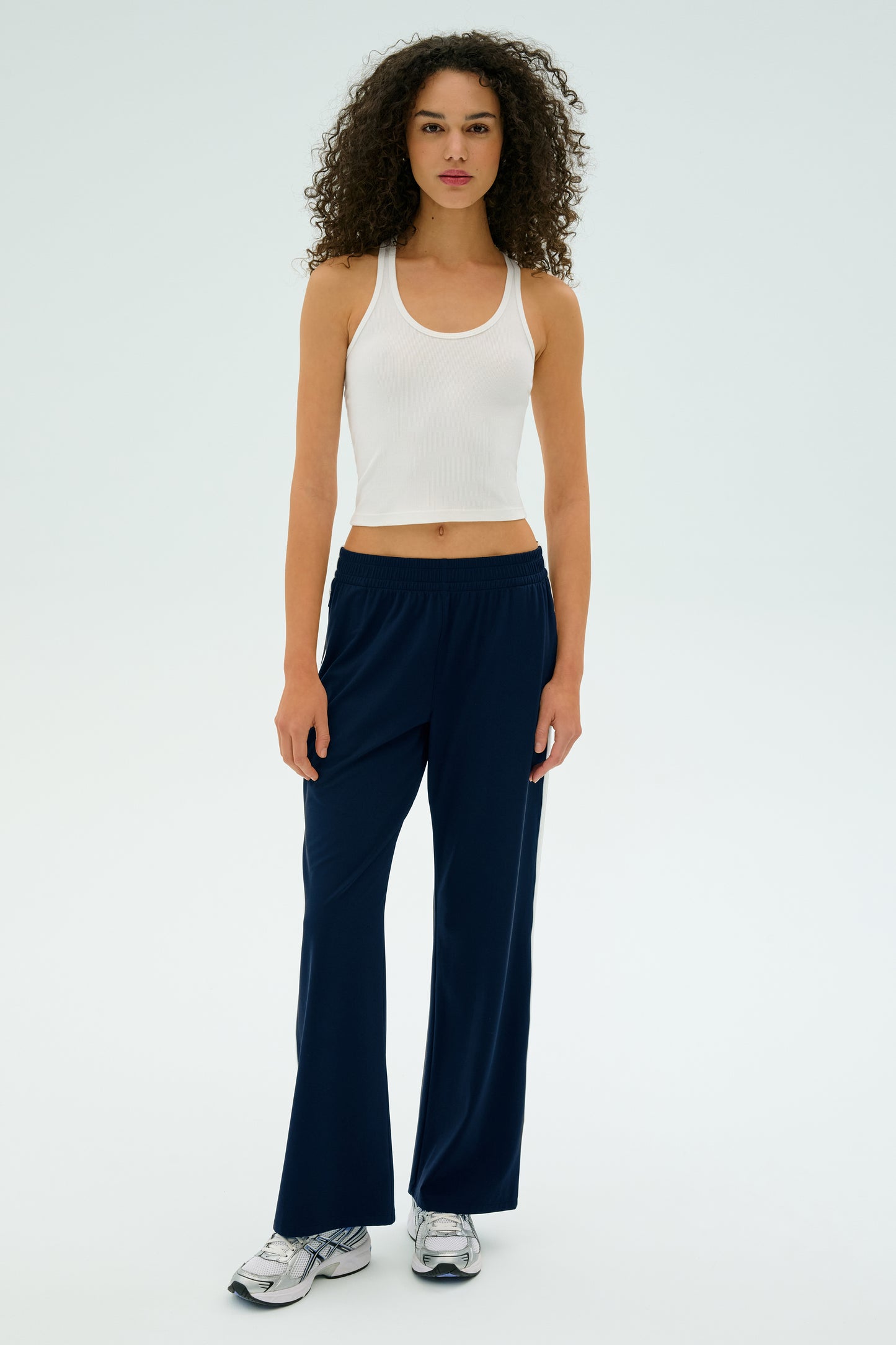 A person with curly hair stands against a light background, wearing a white tank top, SPLITS59's Maxie Rigor Track Pant in Indigo/White, and sneakers.