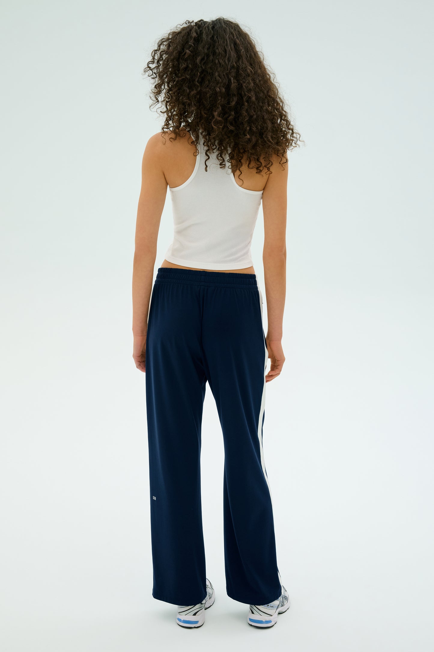 A curly-haired person in a white tank top and Maxie Rigor Track Pant by SPLITS59, featuring a wide leg silhouette, stands facing away against a plain background, paired with sneakers.