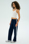 A person with curly hair stands against a plain background, stylishly dressed in a white tank top and SPLITS59's Maxie Rigor Track Pant - Indigo/White featuring a wide-leg navy-blue silhouette with white stripes, effortlessly paired with silver sneakers.