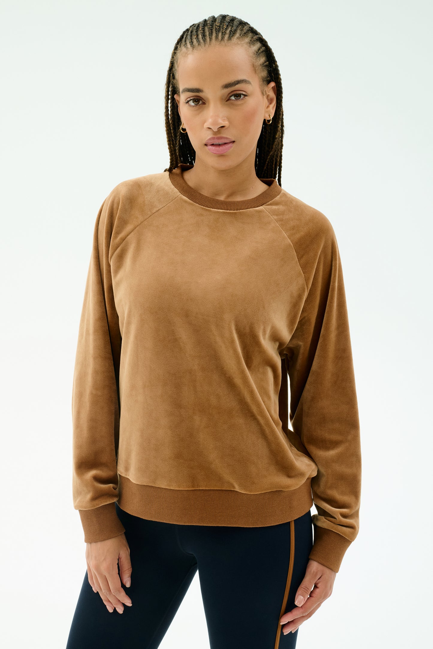 A woman with braided hair wears the SPLITS59 Joy Velour Crewneck in Honey, paired with dark side-striped stretch pants, standing against a plain light background.
